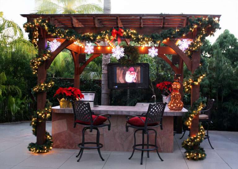 Wood outdoor pergola finished in Early American stain with Champion profile, featuring holiday garland, string lights, an outdoor TV, bar seating, and speakers for backyard entertaining.