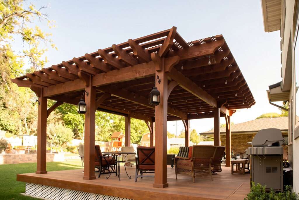Full wrap cantilever roof pergola covering an elevated backyard deck with timber posts, decorative knee braces, outdoor seating, and integrated lighting.