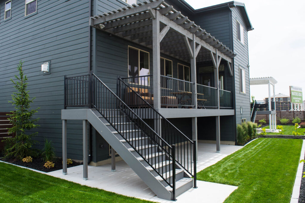 Attached timber frame pergola over elevated backyard deck with black metal railing and stairs. Attached pergola in Canyon Grey stain with open roof casting shadows over a wood dining table on a raised patio deck.