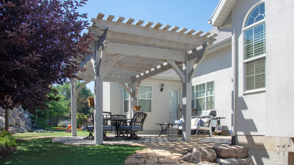 Bleach-stained timber pergola over backyard patio with stone pavers and outdoor dining set, adjacent to white stucco home.