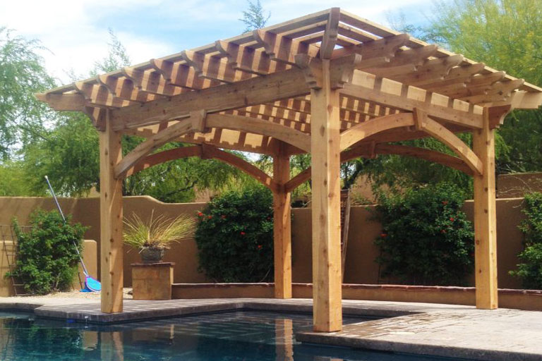 Natural cedar pergola with arched braces and keystones casting shade across a pool step in an Arizona backyard