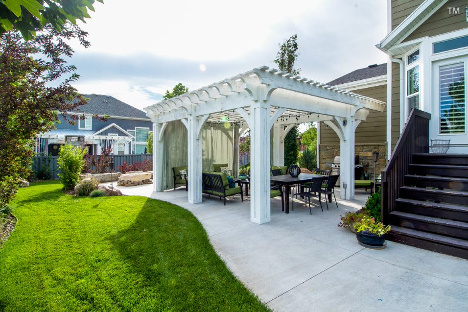 Alpine-stained white timber pergola with cantilevered roof, outdoor curtains, string lights, and patio seating in a landscaped backyard.