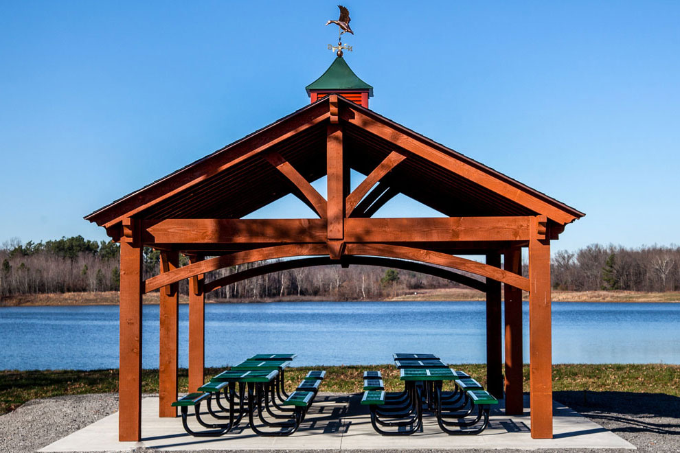 Timber frame pavilion stained in Rich Sequoia with a green metal roof and eagle weathervane, set beside a lake in a Kentucky park.
