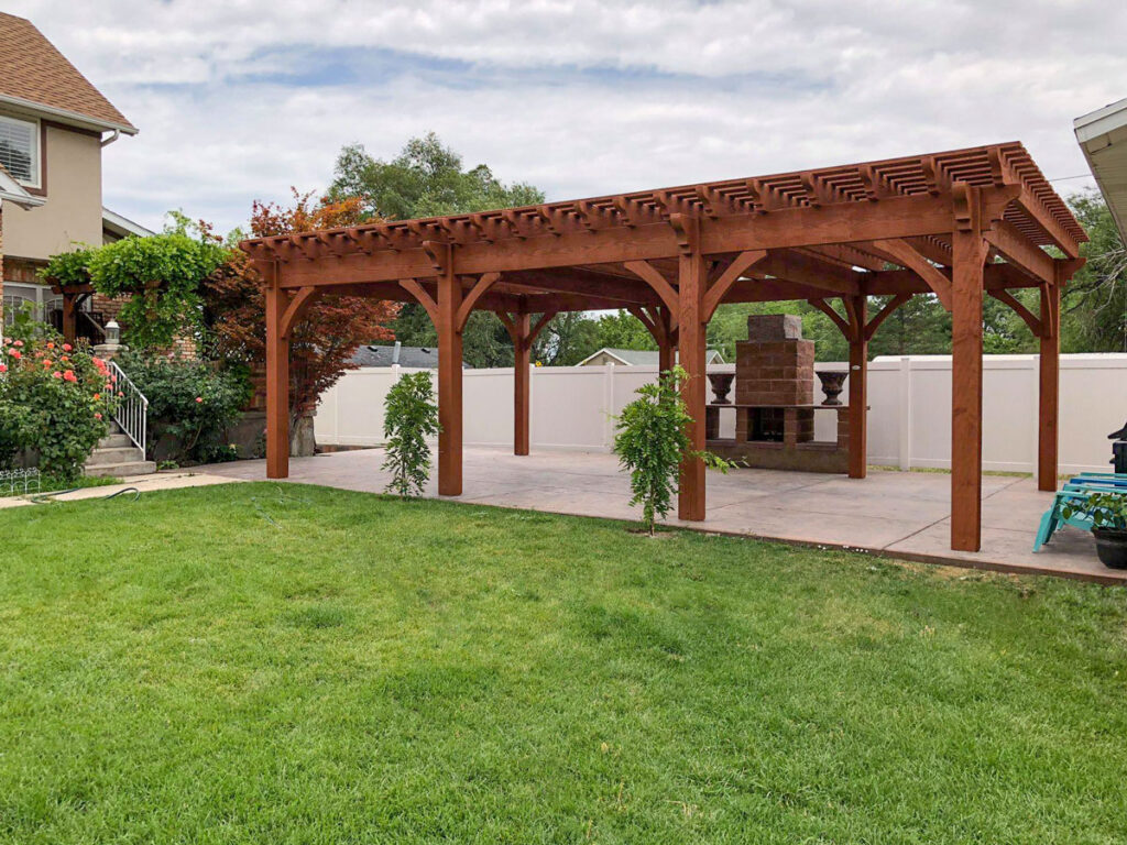 Large timber frame pergola stained in Rich Sequoia, covering an outdoor patio and stone fireplace in a residential backyard.