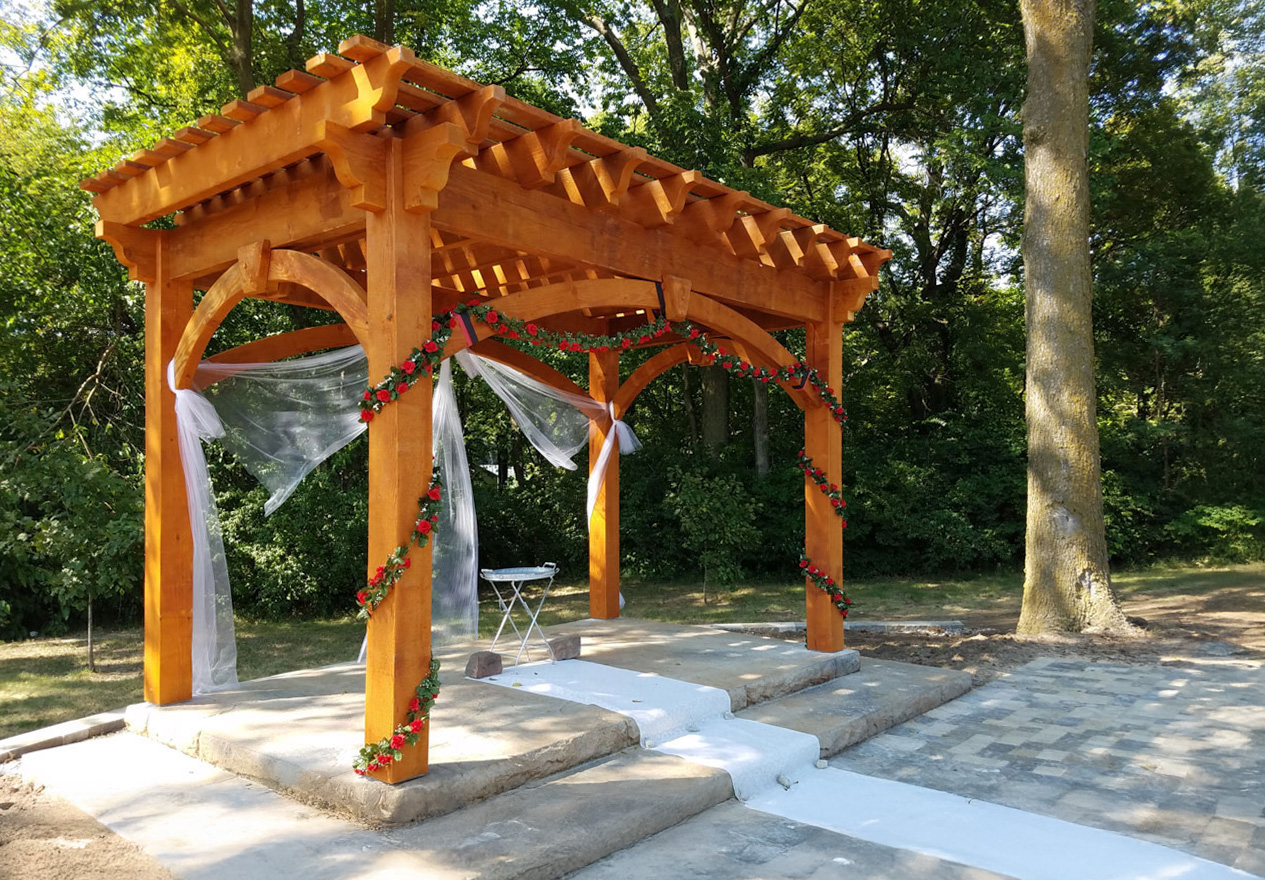 Handcrafted timber wedding pergola with arched beams, draped fabric, and rose garlands in a sunlit woodland setting.