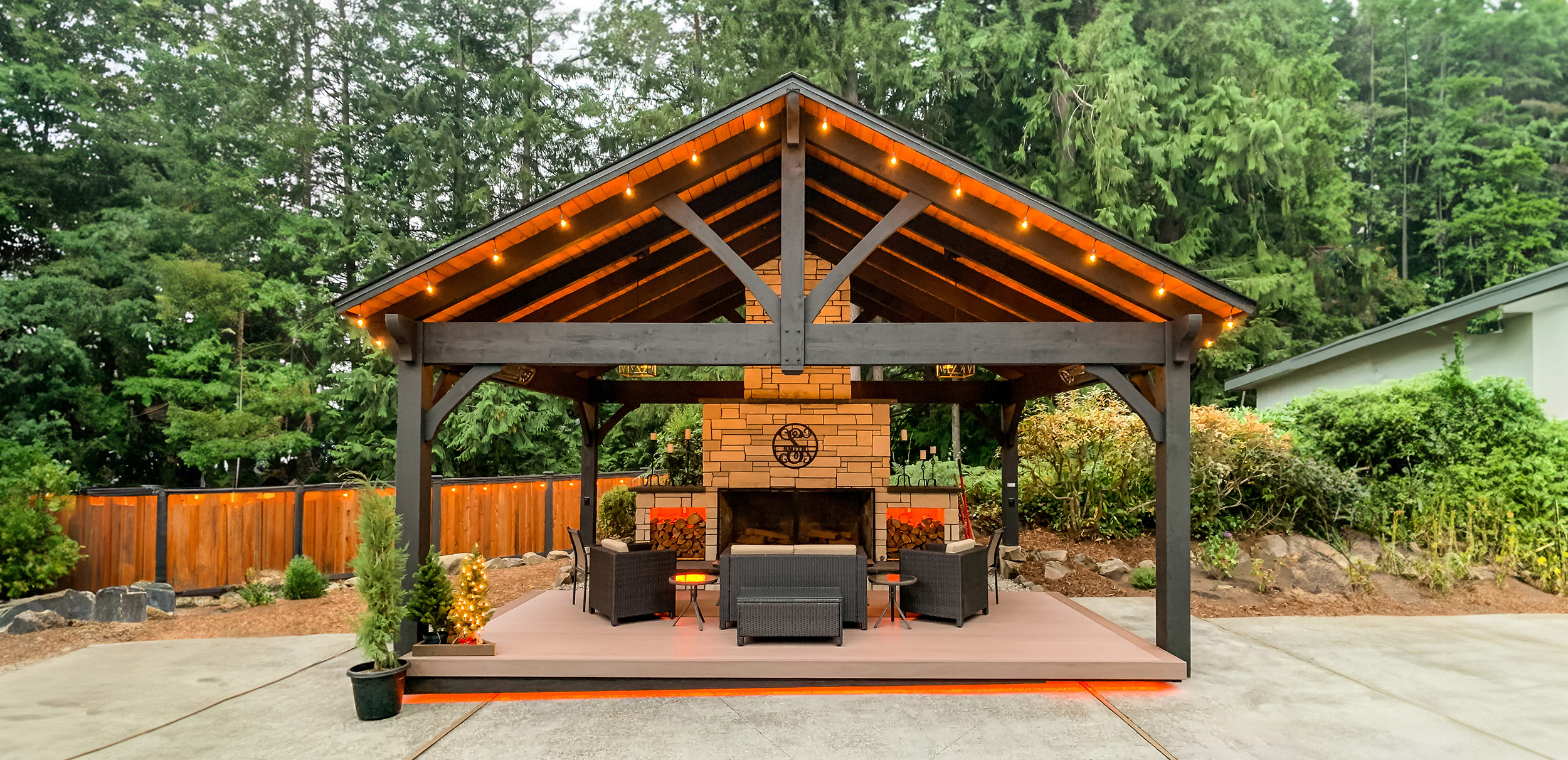 Timber frame pavilion with gable roof and outdoor fireplace, surrounded by string lighting and forest landscaping.
