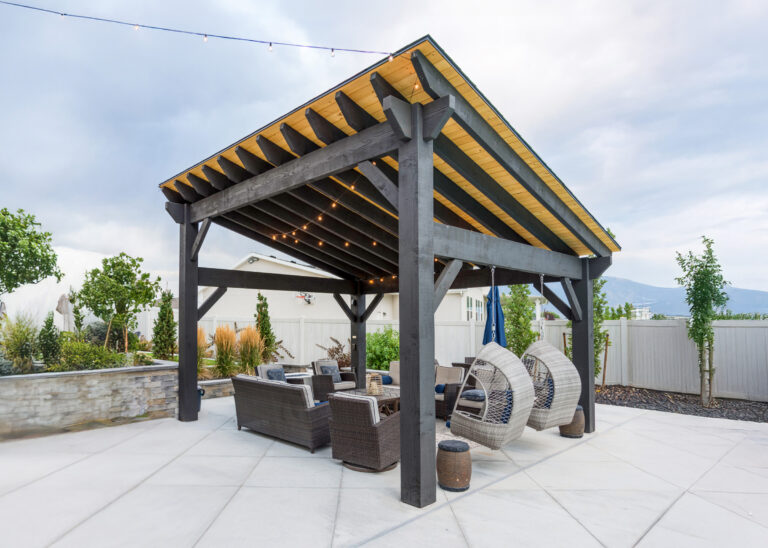 Modern timber frame cabana with a shed-style roof in black ebony stain, featuring cozy poolside seating, hanging wicker chairs, and warm string lights beneath natural tongue-and-groove ceiling panels.