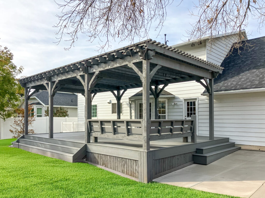 Slate gray timber pergola with built-in bench seating and raised deck, attached to a white house with concrete patio and bright green lawn.