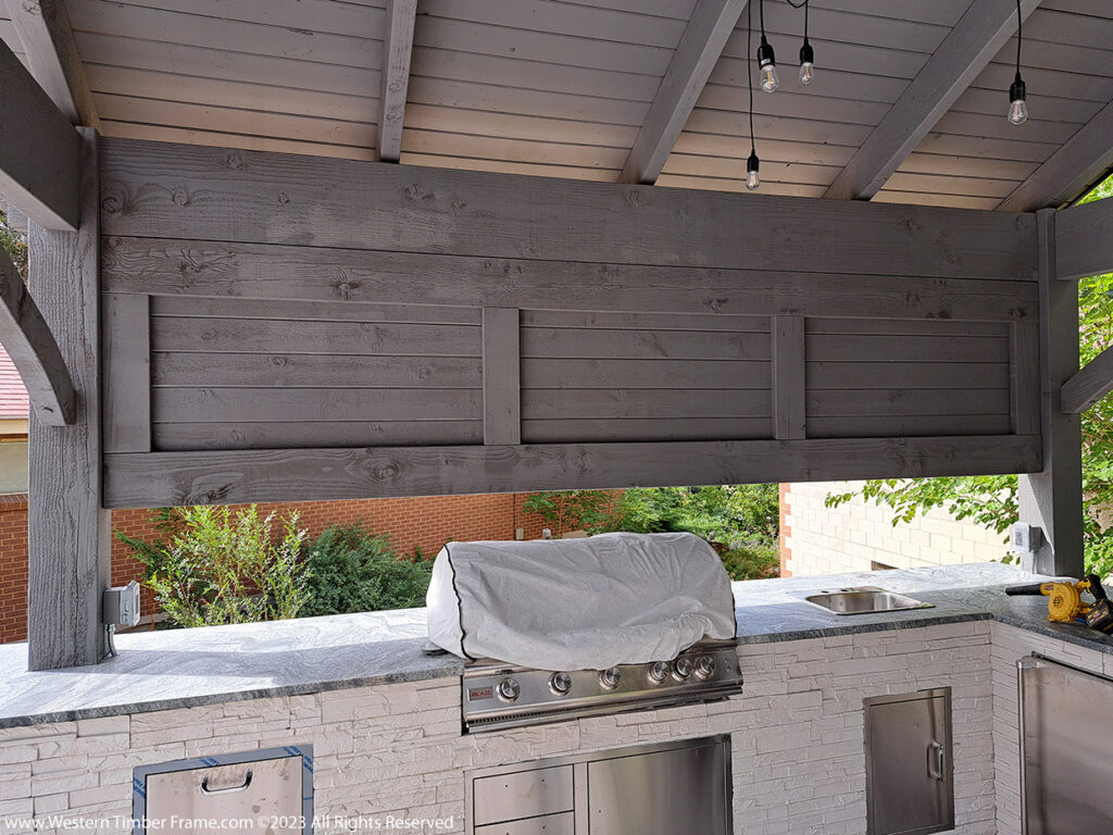 Western Timber Frame™ timber frame pavilion featuring a solid privacy wall stained in gray, partially enclosing a built-in outdoor kitchen with stone counters, stainless steel appliances, and hanging lights.