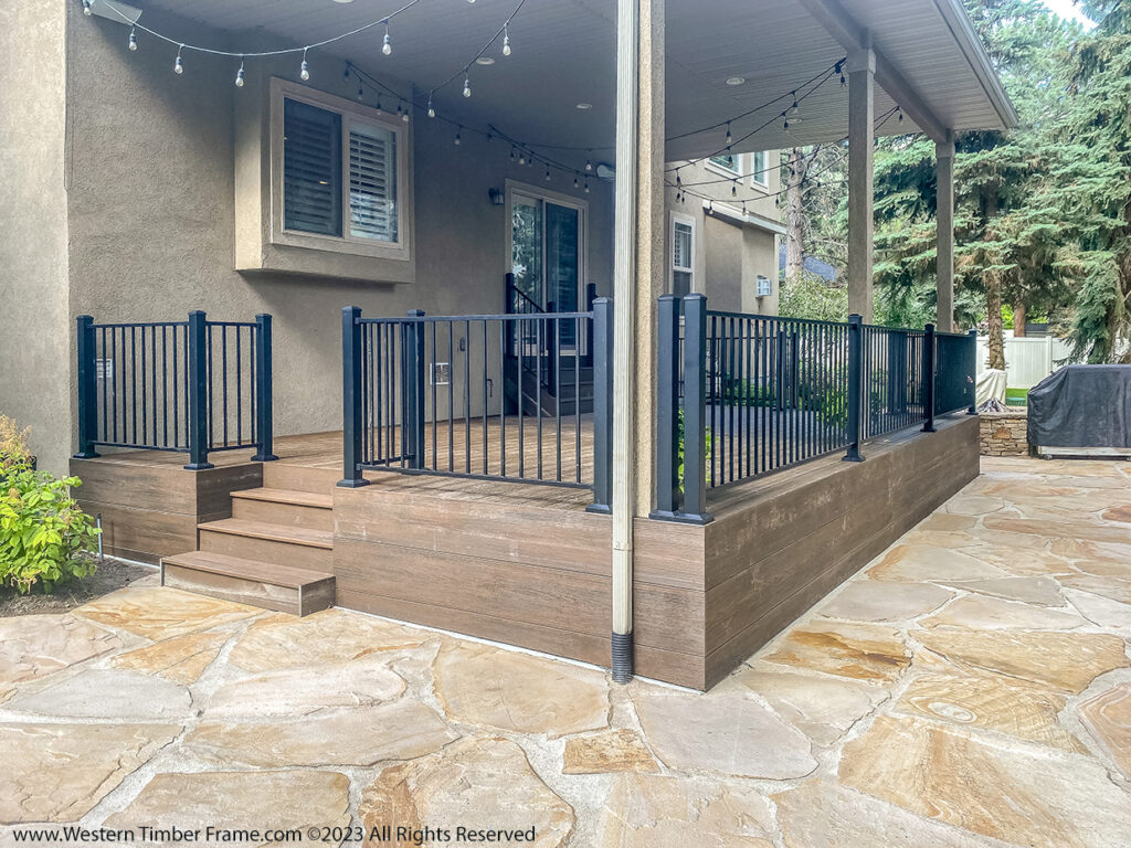 Western Timber Frame™ timber frame deck featuring a solid skirt finish around the sides and black metal railing, connected to a home’s back patio with a flagstone walkway.