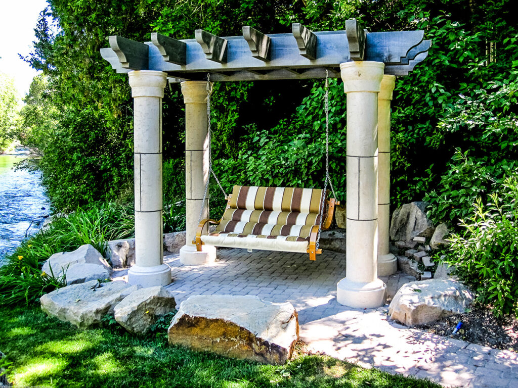 Western Timber Frame™ timber frame garden arbor with dark-stained beams and stone Tuscany-style columns, featuring a striped cushioned bench swing by a scenic stream.