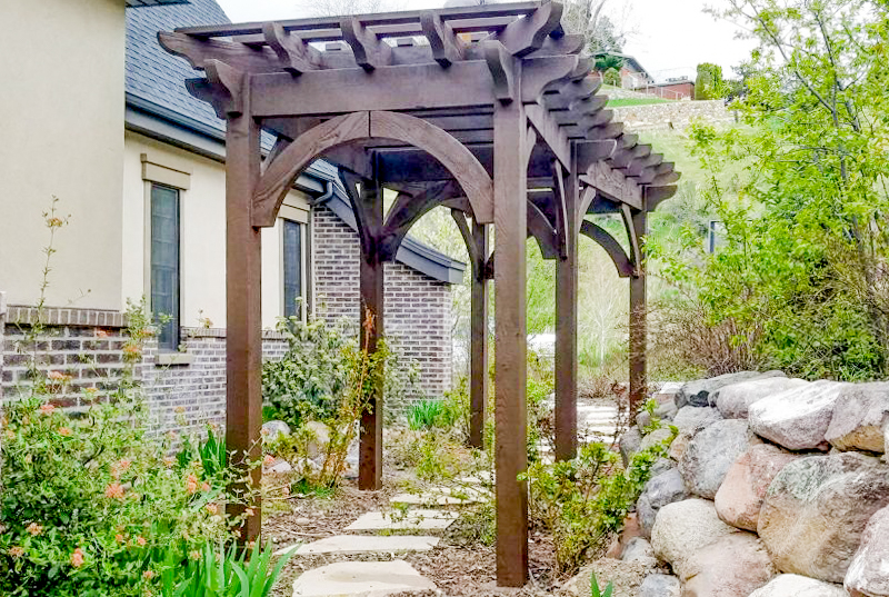 Western Timber Frame™ timber frame arbor with arched knee braces and a dark stain, installed over a flagstone garden pathway next to a brick and stucco home.