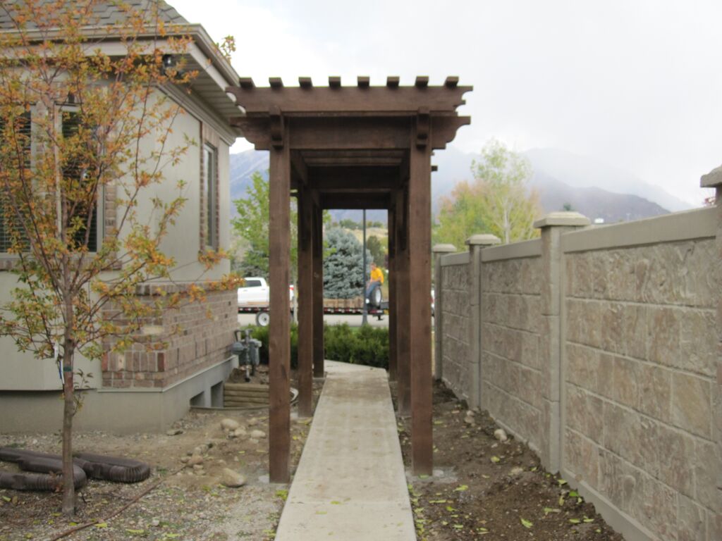 Western Timber Frame™ timber frame arbor with a dark wood finish installed over a concrete walkway alongside a home and privacy fence, set against a backdrop of trees and mountains.