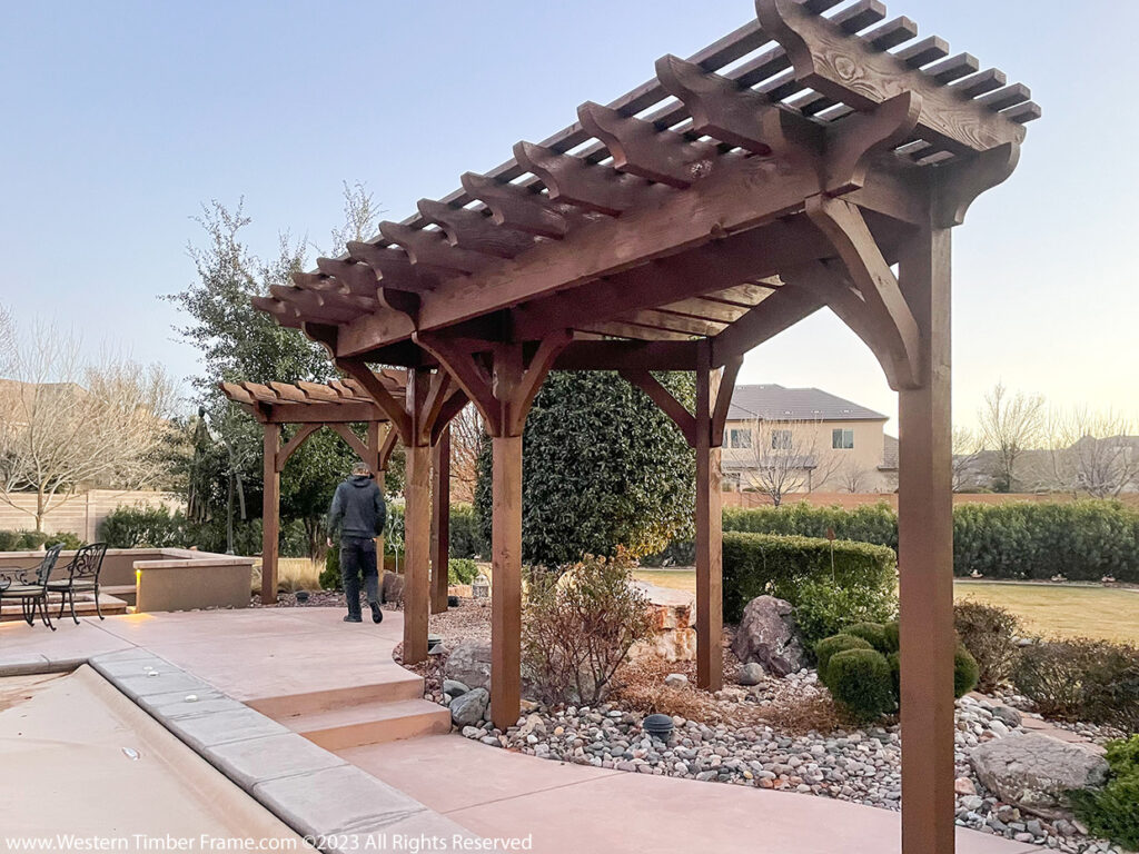 Western Timber Frame™ poolside timber frame pergola and trellis with a rich wood stain, set along a landscaped backyard patio with seating areas and a garden path.