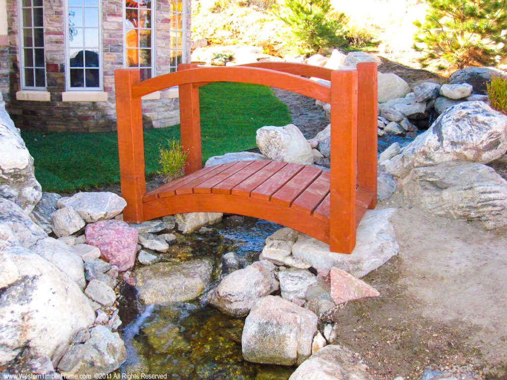 Western Timber Frame™ timber frame garden bridge finished in a rich Sequoia stain, arching gracefully over a natural stream surrounded by rocks and landscaping.