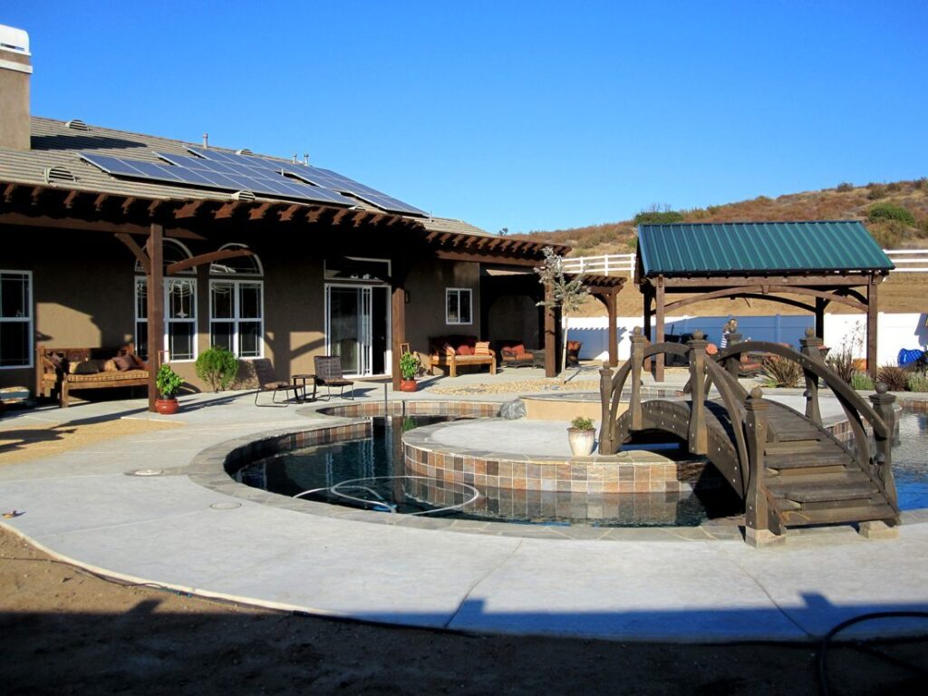 Western Timber Frame™ timber frame pergolas attached to a California home, a freestanding pavilion with a green metal roof, and an arched timber bridge over a custom pool with a spa.