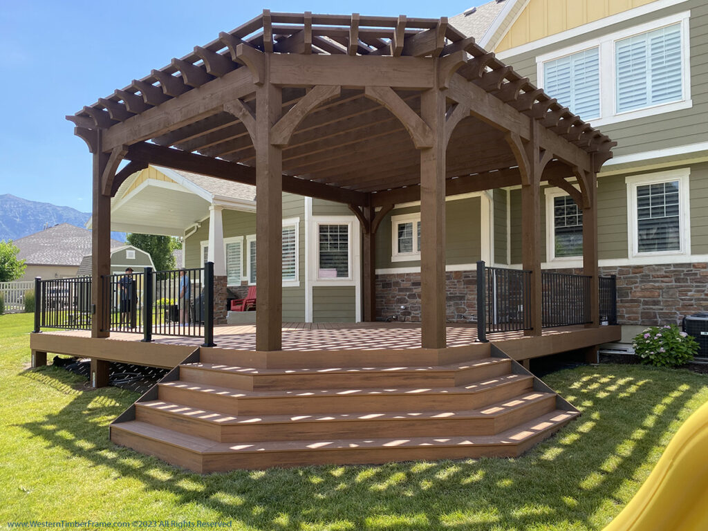 Western Timber Frame™ timber frame pergola with dark wood finish, built over a custom elevated deck featuring black metal railing and wide wraparound steps in a landscaped backyard.