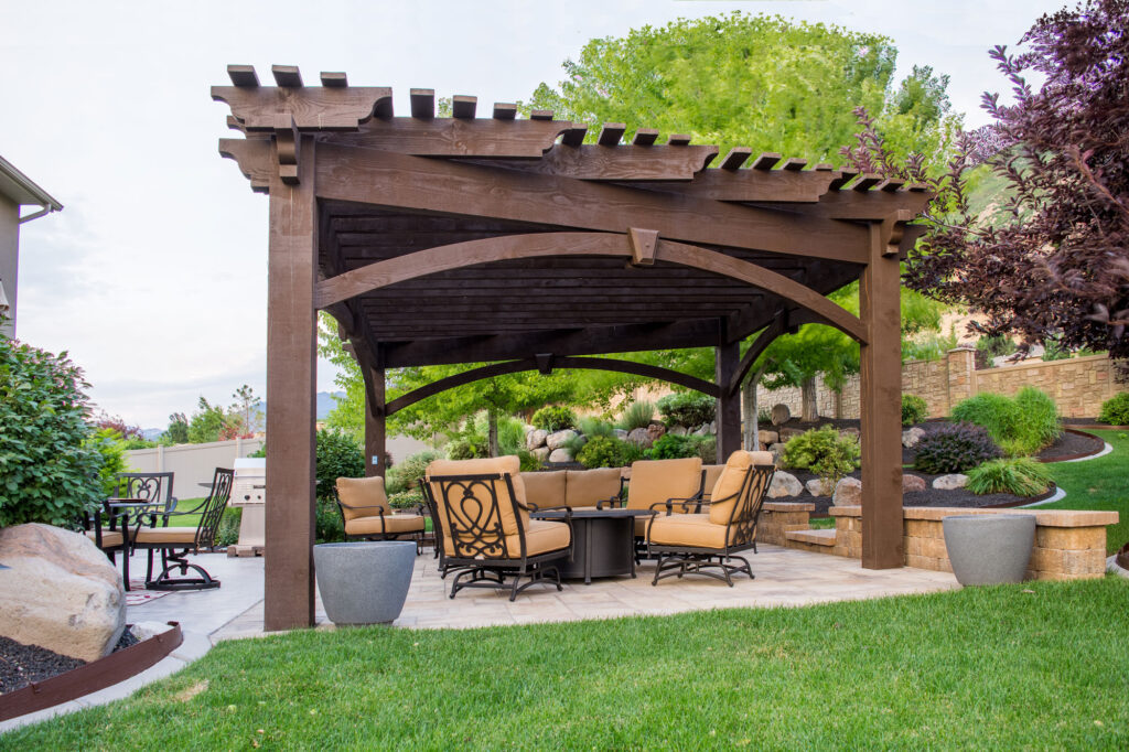 Western Timber Frame™ custom-shaped timber frame pergola with dark stain and a TimberVolt® power post, covering a comfortable outdoor seating area in a beautifully landscaped backyard.