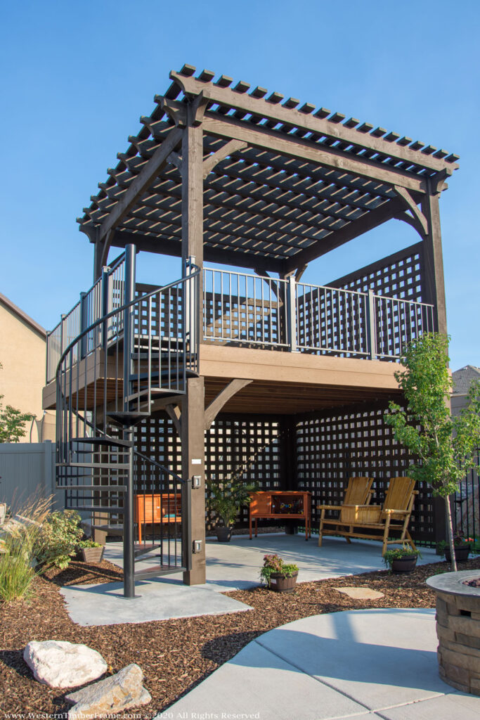Western Timber Frame™ timber frame two-story pergola with dark stain, latticed privacy walls, a black spiral staircase, and integrated TimberVolt® power post in a landscaped backyard.