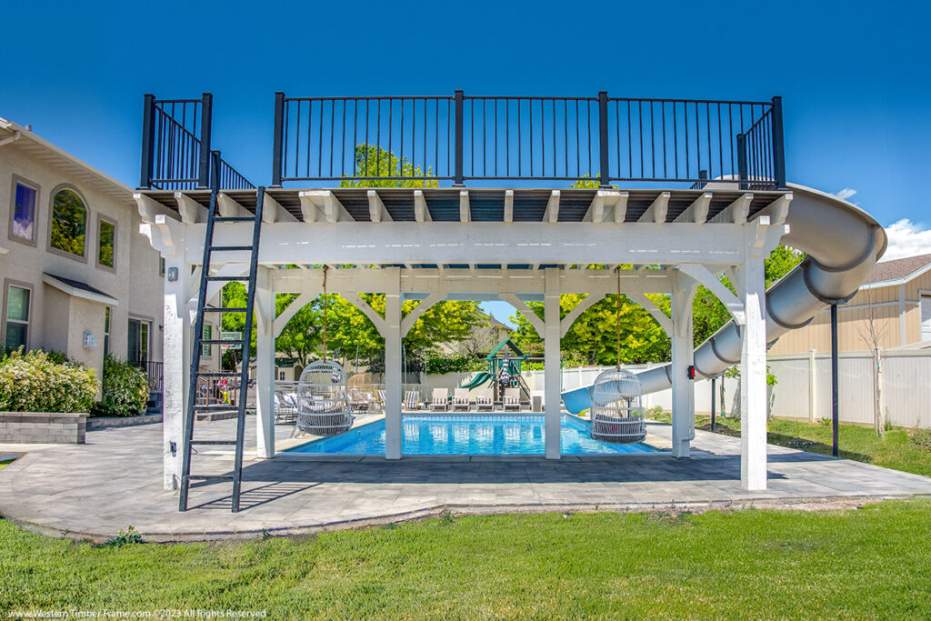 Western Timber Frame™ white timber frame pergola with an elevated diving deck, TimberVolt® power posts, a built-in twisting slide, and two hanging egg swings over a backyard pool.