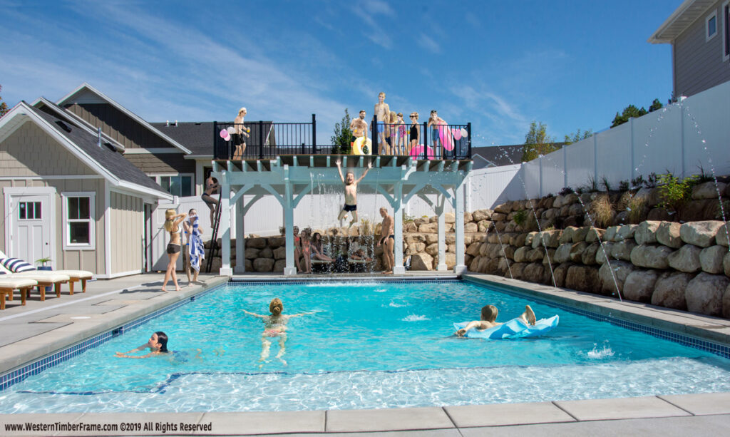 Western Timber Frame™ timber frame poolside pergolas with a diving deck and waterfall feature, TimberVolt® power posts, and people enjoying swimming, diving, and lounging around a backyard pool.