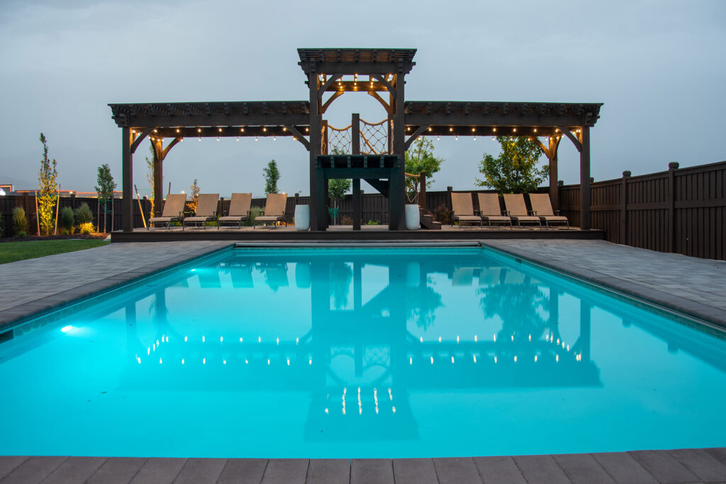 Western Timber Frame™ timber frame poolside step roof pergolas with integrated TimberVolt® power posts, ambient string lights, and a central elevated diving deck, illuminated at dusk.