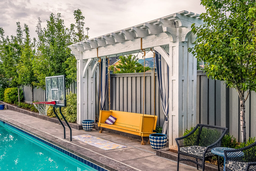 Western Timber Frame™ Alpine White timber frame arbor installed beside a swimming pool, featuring a vibrant yellow bench swing suspended with rope and accented with potted plants and lounge chairs.