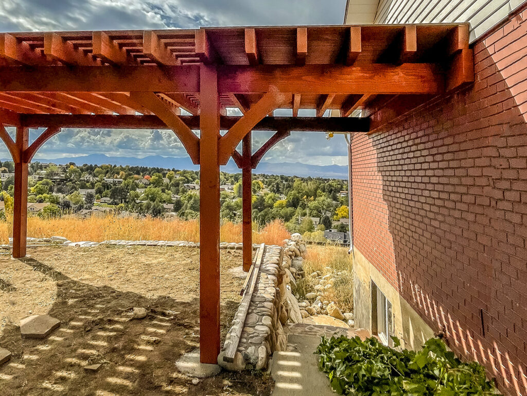 Western Timber Frame™ timber frame pergola and pavilion extension providing shade and protection over a basement entryway, set against a scenic hillside view.