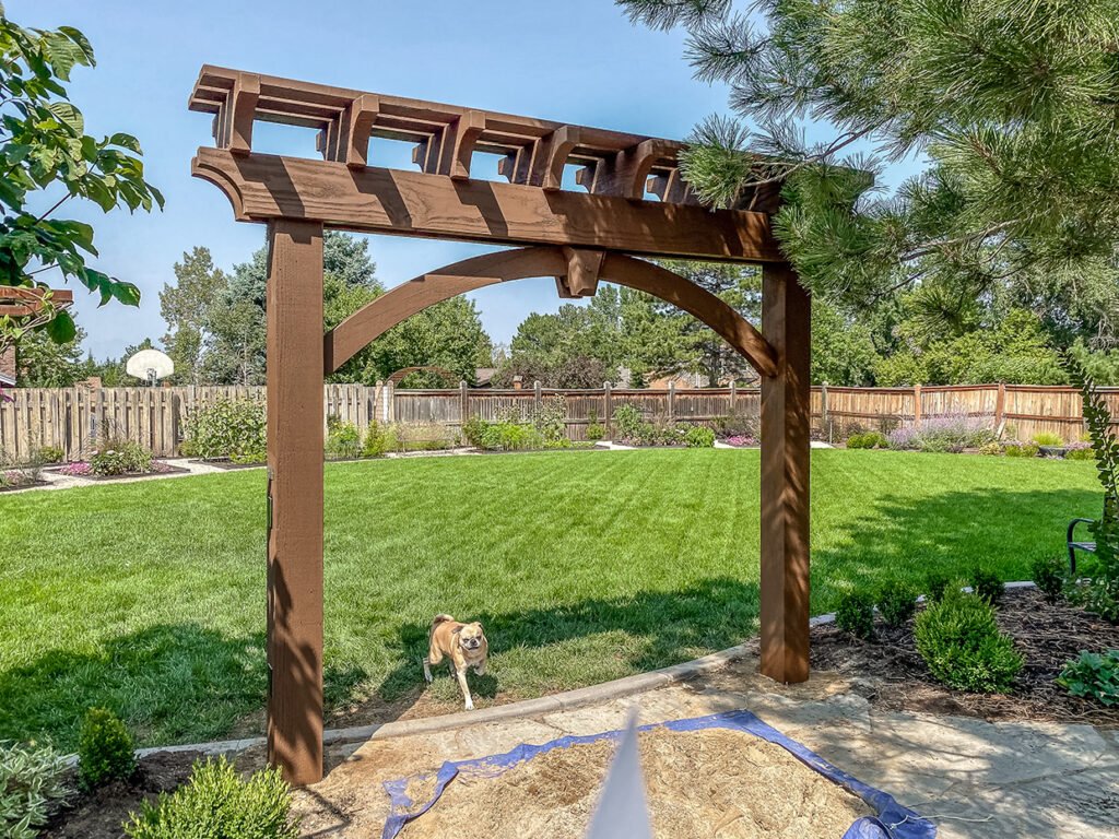 Timber frame garden arbor by Western Timber Frame™ featuring decorative beam work in a landscaped backyard with a dog running through.