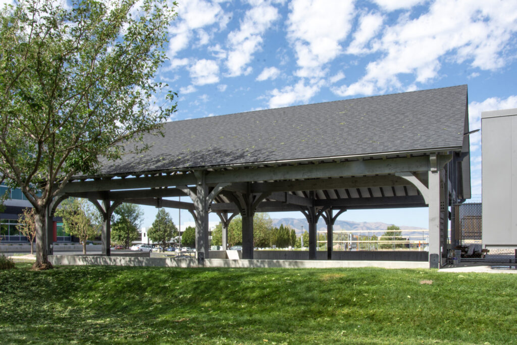 Large timber pavilion stained in Canyon Grey with pitched roof, open sides, and surrounding green space in a public park.