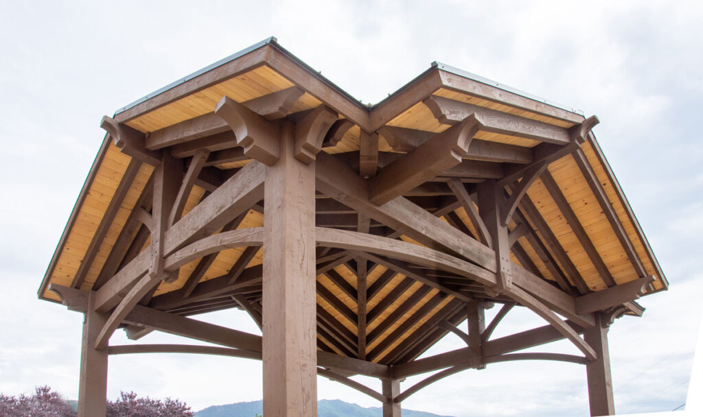 Timber frame pavilion with cross-beam gabled roof featuring natural wood stain and dark brown contrast by Western Timber Frame