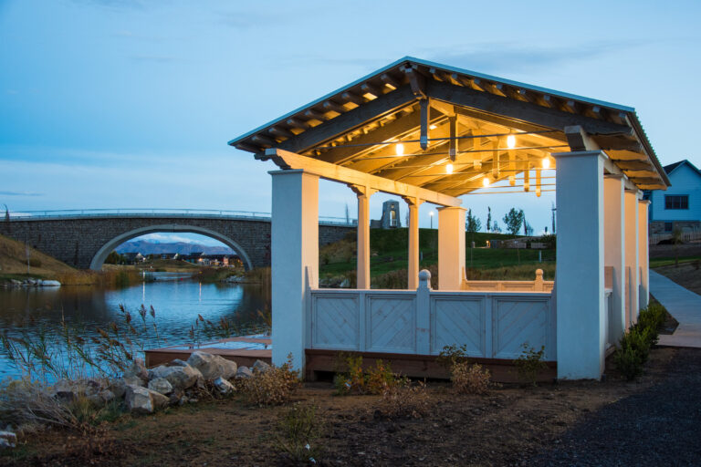 Illuminated timber frame pavilion built by Western Timber Frame in Daybreak, Utah, overlooking a calm lake with a stone bridge and mountain views at dusk.