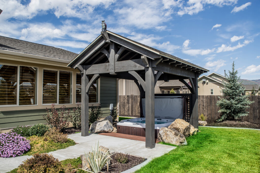 Small timber frame pavilion stained in Canyon Grey, covering a hot tub in a landscaped backyard with stone accents.