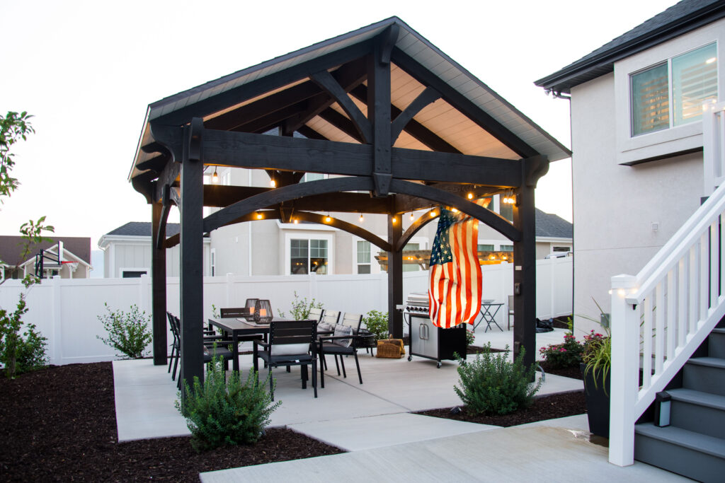 Ebony-stained timber pavilion with pitched roof and American flag, set over a concrete patio with outdoor dining and string lights.