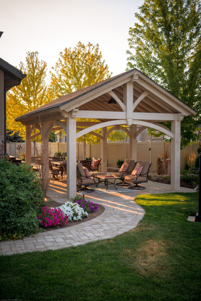 Custom-stained timber frame pavilion with arched trusses and cozy outdoor seating, adjacent to a stucco home.