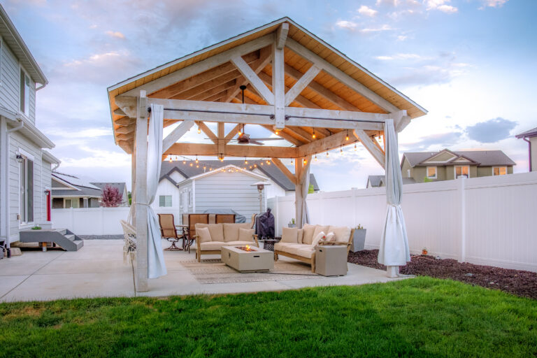 Bleach-stained timber pavilion with gable roof, outdoor string lights, and cozy seating area with fire pit and curtains.