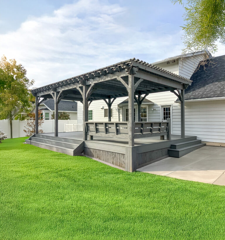 Slate gray timber pergola with built-in bench seating and raised deck, attached to a white house with concrete patio and bright green lawn.