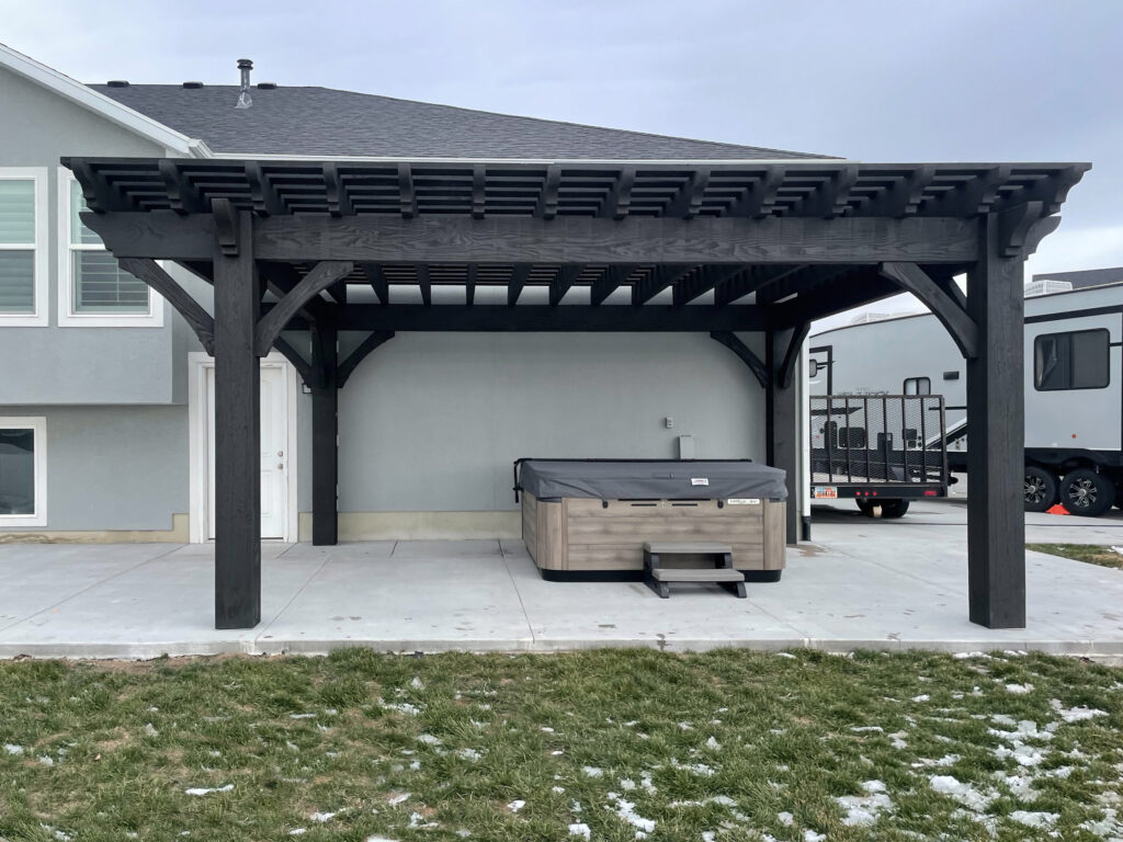 Large timber pergola in deep Ebony stain shading a hot tub on a backyard concrete patio, set against a light gray home exterior.