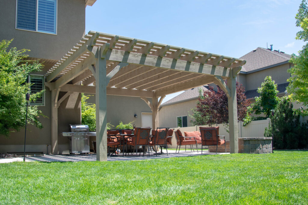 Backyard timber frame pergola stained in Beach Sand, shading an outdoor dining set and grill station beside a stucco home.