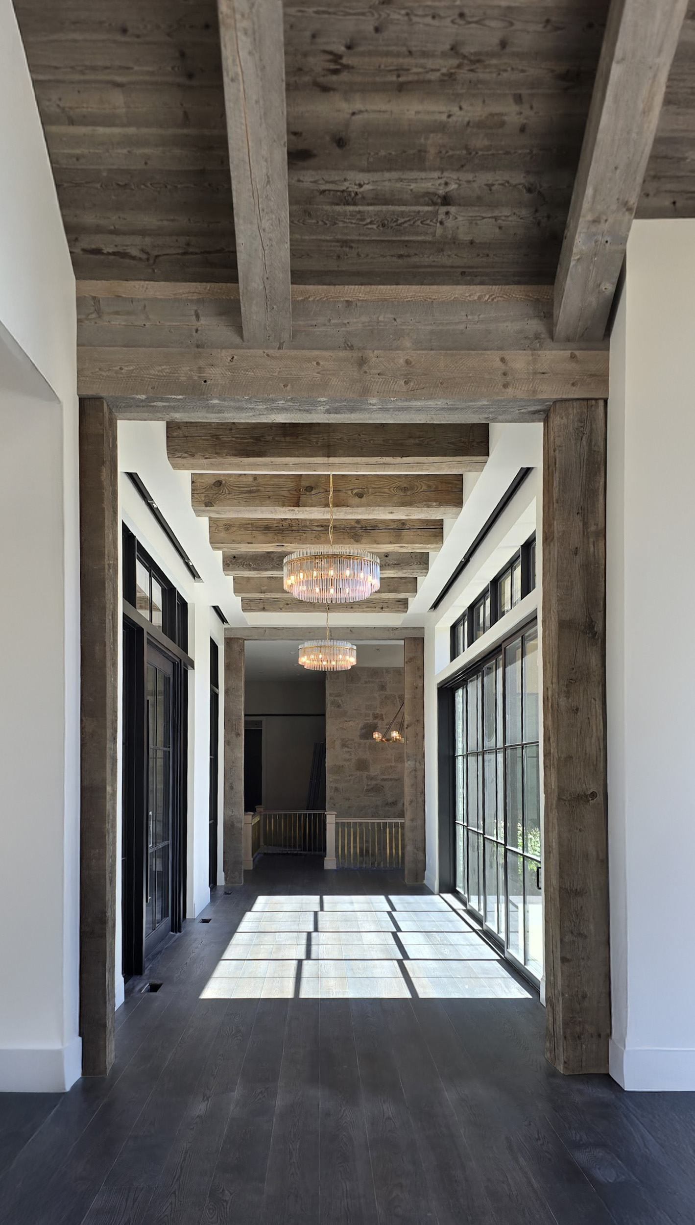 Exposed timber ceiling with beams and posts by Western Timber Frame™ in a luxury hallway with chandeliers and tall black-framed windows
