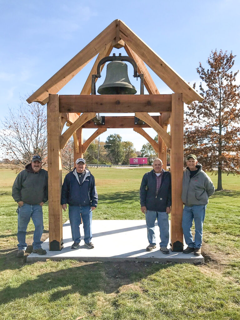 Four men pose beside a handcrafted timber frame bell structure by Western Timber Frame™ in Indiana, designed for strength, beauty, and lasting legacy