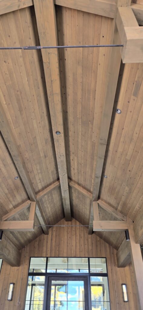 Looking upward into the interior of a Western Timber Frame™ structure, this image showcases a soaring timber frame ceiling with exposed beams and precision-fitted joinery. The tongue and groove wood ceiling adds warmth and texture, drawing the eye along the natural grain and craftsmanship. Rich wood tones, soft lighting, and the strength of architectural-grade timbers create a sense of grounded elegance and alpine retreat.