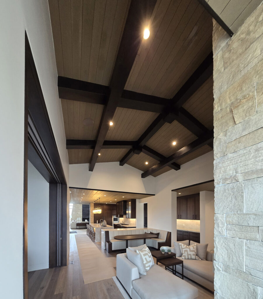 Handcrafted exposed timber frame and tongue-and-groove ceiling with elegant lighting, creating a warm, luxurious atmosphere above the kitchen and living room, by Western Timber Frame™
