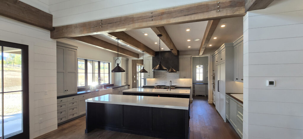 Exposed timber support beam by Western Timber Frame™ in a bright white kitchen with upper loft, modern black railings, and shaker cabinetry.