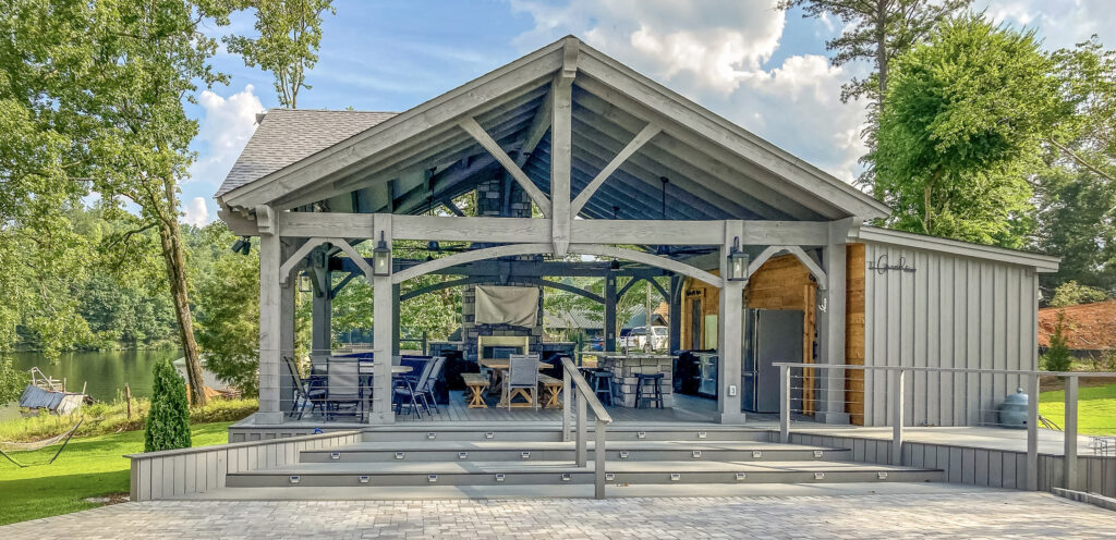 Timber pavilion with a gabled roof, dining area, and outdoor kitchen beside a lake, with light grey stain and modern railing.