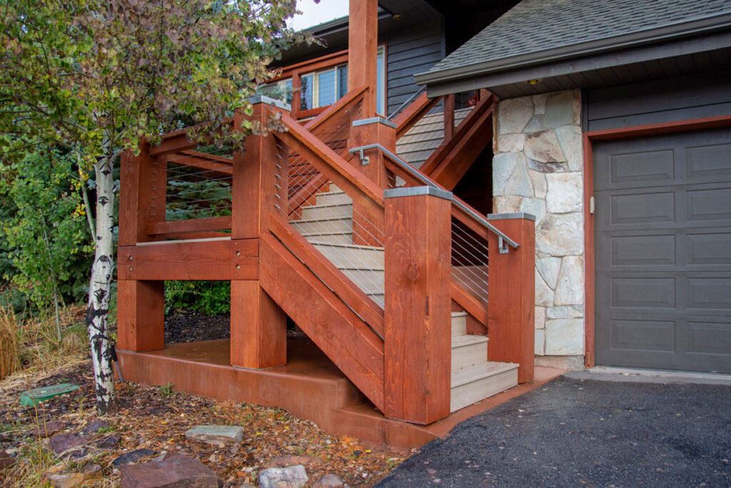Exterior timber stair railing stained in Rich Sequoia with stainless steel cable infill, mounted to a home with stone and lap siding.