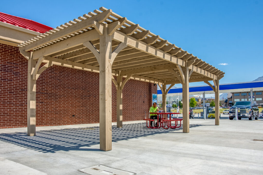Timber frame pergola in Beach Sand stain beside a red brick commercial building, providing shade over a red picnic table on a concrete patio.