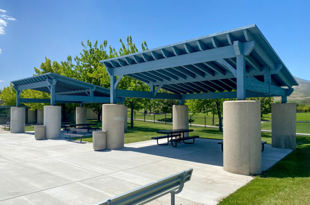 Commercial-grade timber frame pavilions with blue shed-style roofs and concrete columns, installed at a public park for shaded gathering space.