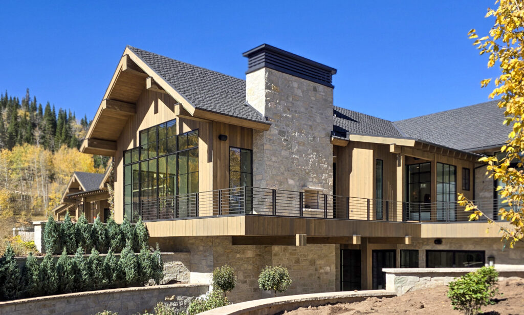 Modern timber mountain home with stone chimney, vertical wood siding, and black steel accents set against an alpine backdrop