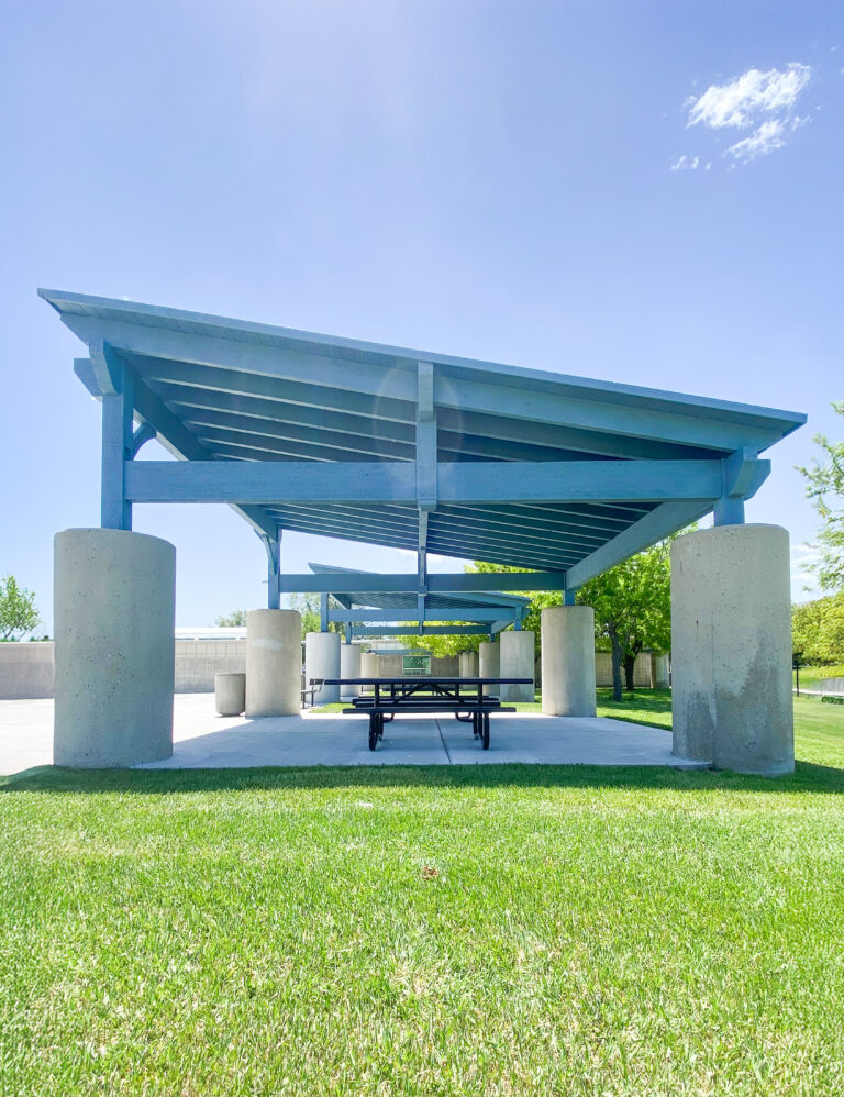 Frontal view of timber frame cabanas with angled shed-style roofs supported by round concrete columns. A picnic table sits beneath the structure on a concrete pad, surrounded by green grass and clear skies.